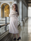 Woman wrapped in a white fur coat standing in an elegant interior setting.