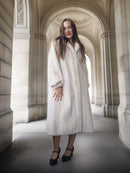 Woman in a long white coat standing in an archway with classical architecture.