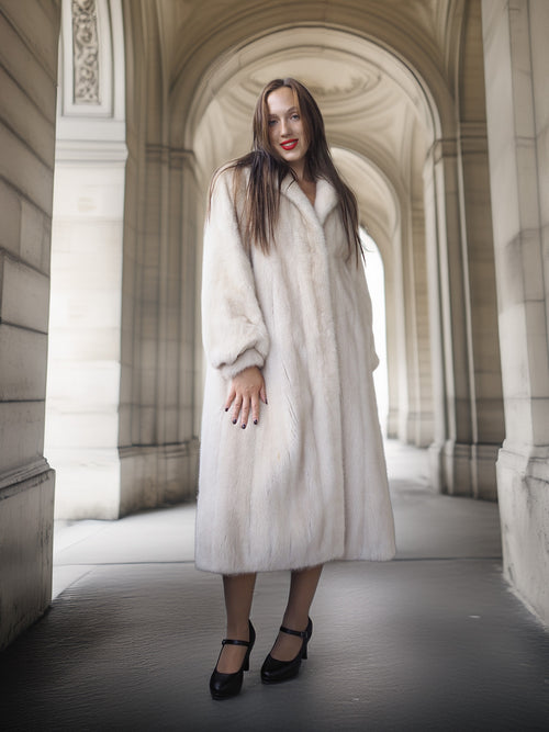 Woman in a long white coat standing in an archway with classical architecture.
