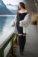 A woman stands on a lakeside path wearing a black off-the-shoulder dress, a voluminous white fur coat, and tall black over-the-knee boots, with mountains and water in the background.