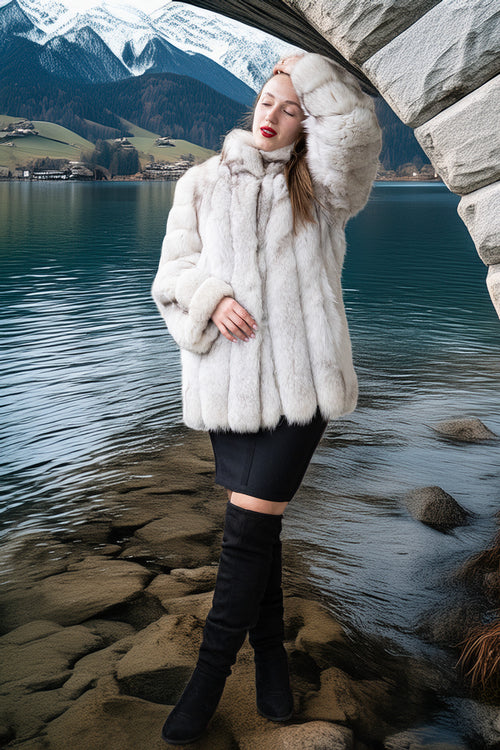 A woman posing in a luxurious white fur coat, black mini skirt, and black over-the-knee boots, standing on rocks by a lake with mountains in the background.