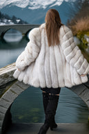 A woman viewed from behind wearing a voluminous white fur coat with quilted paneling, standing on a stone bridge over water with mountains in the background.