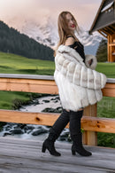 A woman stands on a wooden deck in front of a mountain lodge, wearing a voluminous white fur coat over a black outfit with black knee-high boots.