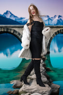 A woman stands on rocks in front of a stone bridge over turquoise water, wearing a black strapless dress, a white fur coat, and black over-the-knee boots.