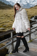 A woman wearing a voluminous white fur coat, black skirt, and black over-the-knee boots stands on a wooden boardwalk in a mountain meadow landscape.