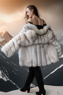 A woman wearing a voluminous white and light gray fur coat with a large plush collar, styled over a black top and black boots, posed against a dramatic mountain backdrop.