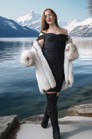 A woman stands by a lake with snow-capped mountains in the background, wearing a black off-the-shoulder mini dress, a white fur coat draped over her shoulders, and black over-the-knee boots.