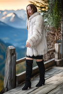 A woman wearing a voluminous white fur coat, black mini skirt, and black over-the-knee boots stands on a wooden deck with a mountain landscape in the background.