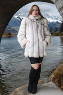 A woman stands outdoors by a lake under a stone bridge, wearing a voluminous white fur coat with a high padded collar, a short black skirt, and black over-the-knee boots. Snow-capped mountains are visible in the background.