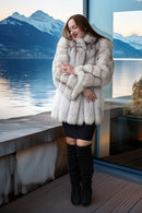 A woman wearing a voluminous light beige fur coat, black mini skirt, and black over-the-knee boots, standing on a wooden deck with a scenic mountain and lake background.