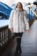 A woman wearing a voluminous off-white fur coat, black mini skirt, and black over-the-knee boots stands on a wooden boardwalk with mountains and snow in the background.