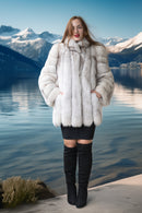 A woman stands outdoors by a lake with snowy mountains in the background, wearing a voluminous white fur coat over a black mini dress and black over-the-knee boots.