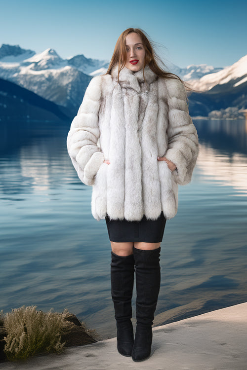 A woman stands outdoors by a lake with snowy mountains in the background, wearing a voluminous white fur coat over a black mini dress and black over-the-knee boots.