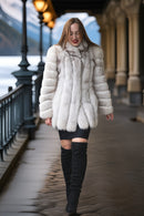 A woman walking on an outdoor covered walkway wearing a voluminous off-white fur coat, a black mini dress, and black over-the-knee boots.