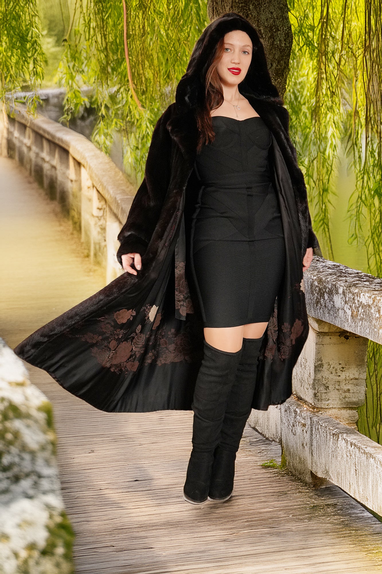 Woman in a black coat and dress standing on a wooden bridge with greenery in the background