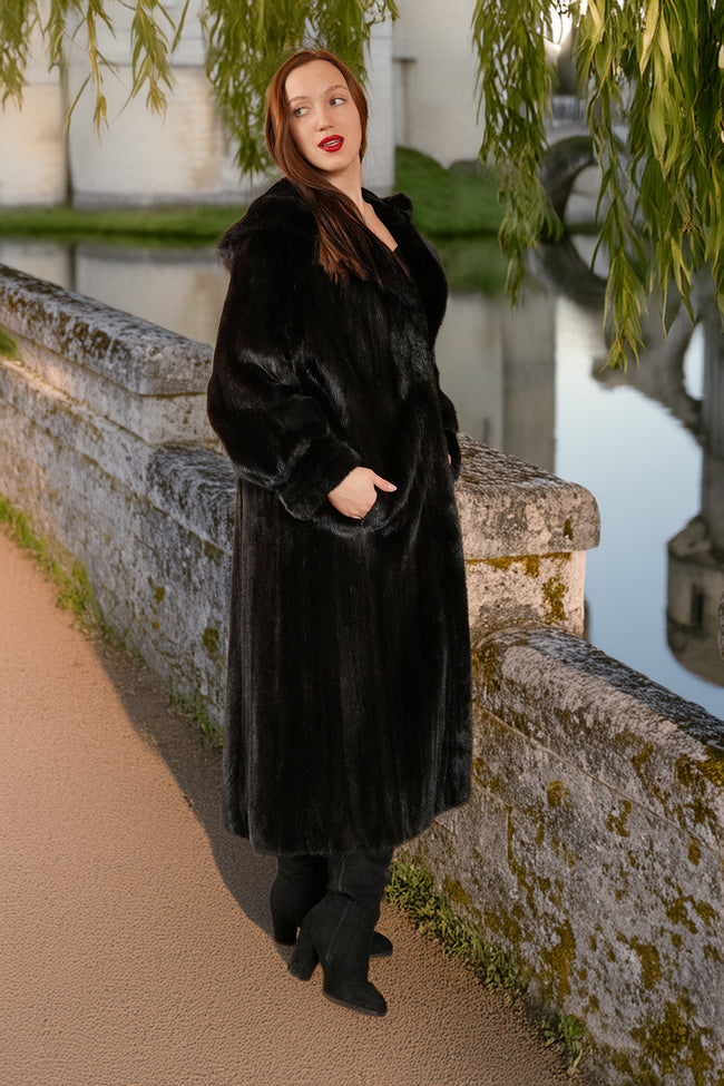 Woman in a black fur coat standing on a stone bridge by a body of water.