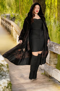 Woman in a black coat and dress standing on a wooden bridge with greenery in the background