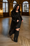 Woman in a long black fur coat standing in an elegant room with wooden floors and large windows.