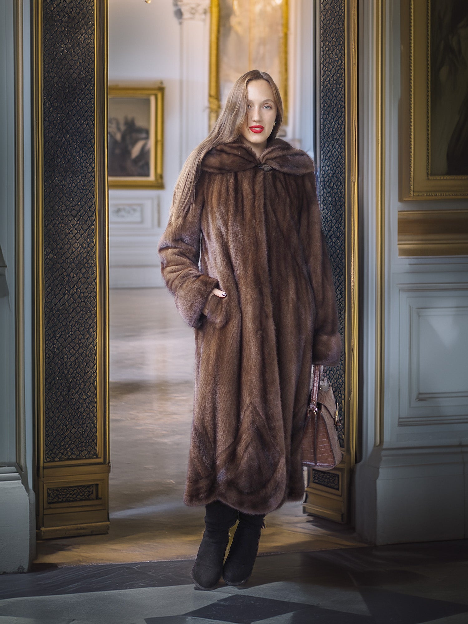 Woman in a long brown fur coat standing in an elegant interior setting.