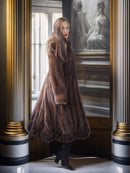Woman in a fur coat standing in a room with classical architecture and a painting in the background.