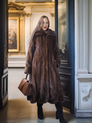 Woman in a long brown fur coat standing in an elegant interior setting.