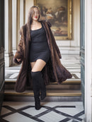 Woman wearing a long brown fur coat, black dress, and knee-high boots in an elegant interior setting.