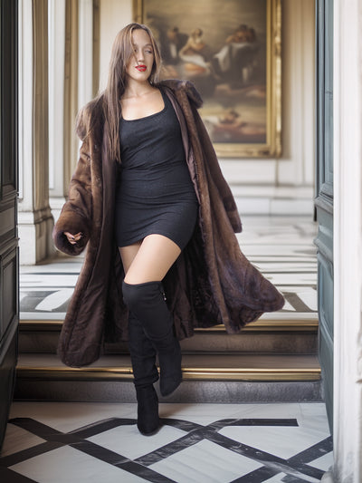 Woman wearing a long brown fur coat, black dress, and knee-high boots in an elegant interior setting.