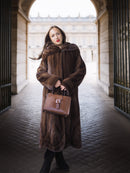 Woman in a long brown fur coat holding a matching handbag in an architectural setting.