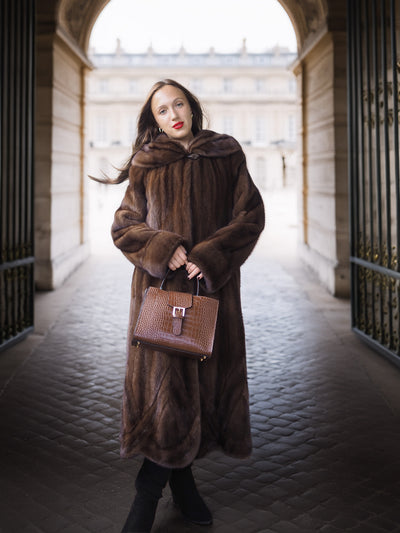 Woman in a long brown fur coat holding a matching handbag in an architectural setting.