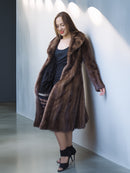 Woman wearing a long brown fur coat against a white wall.