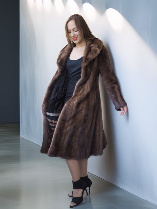 Woman wearing a long brown fur coat against a white wall.