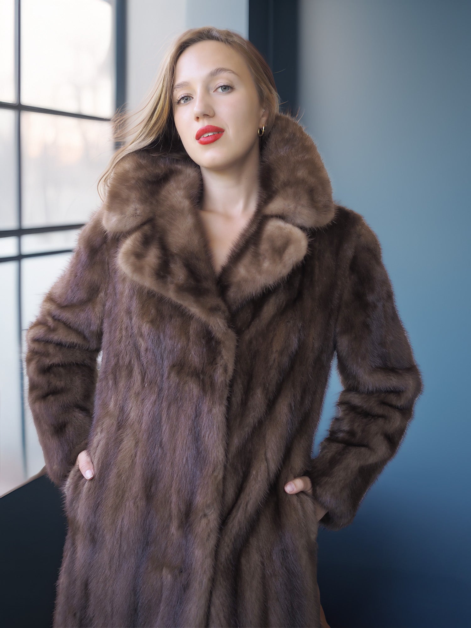 Woman wearing a brown fur coat in a room with large windows.