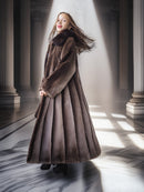 Woman in a long brown coat standing in a sunlit hallway with columns.