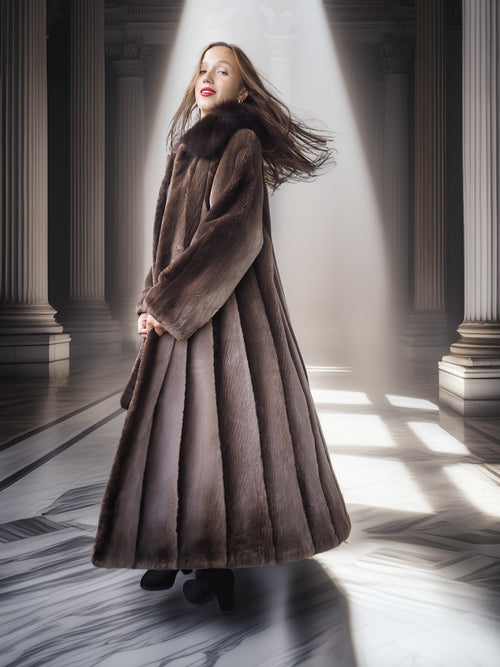 Woman in a long brown coat standing in a sunlit hallway with columns.