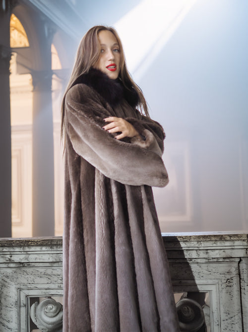 Woman wearing a long brown fur coat standing indoors with architectural elements in the background.
