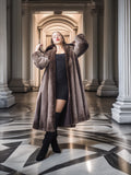Person wearing a long brown coat in an elegant interior setting with columns and a patterned floor.
