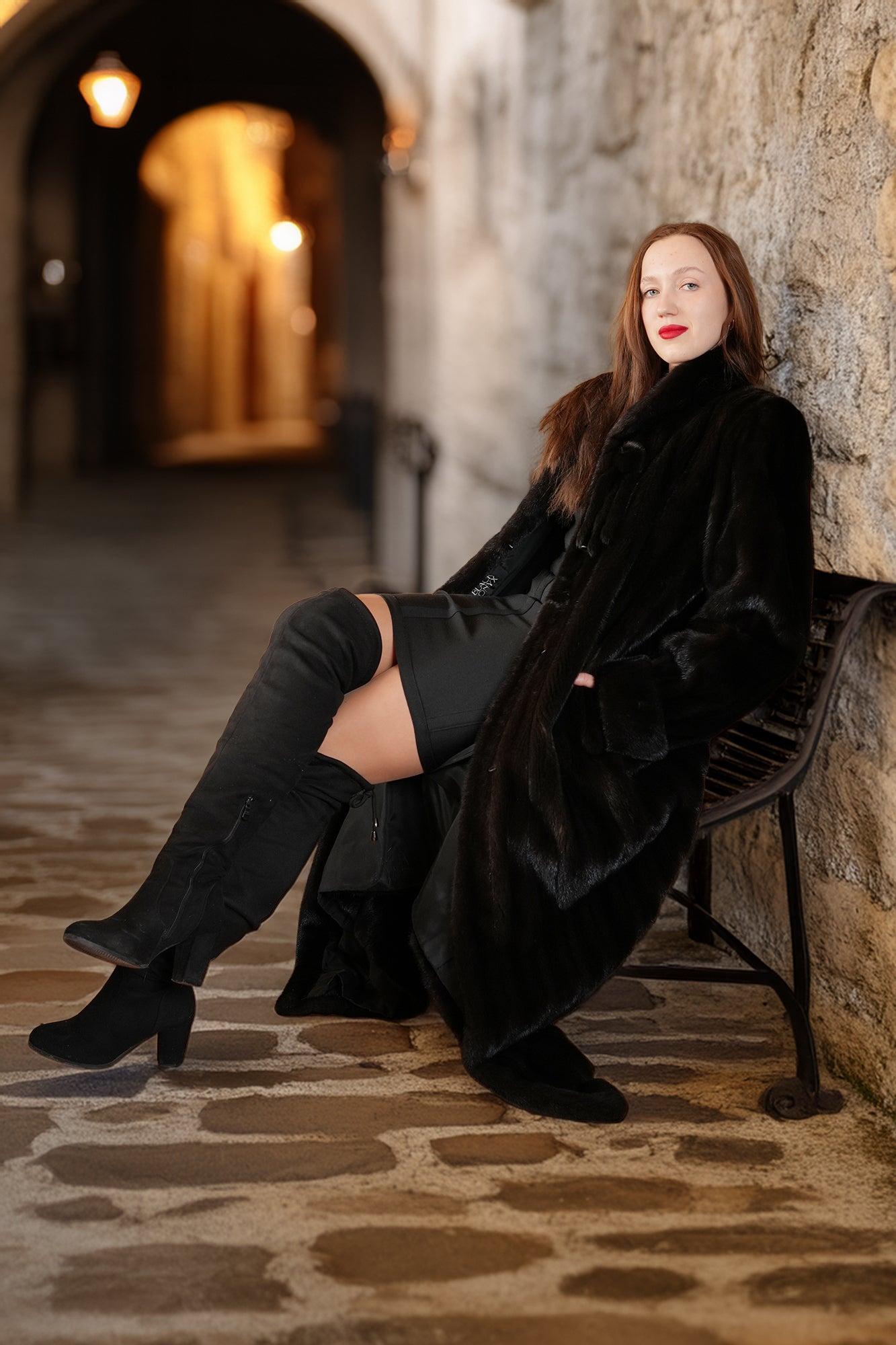 Woman in a black coat and boots sitting against a stone wall in a dimly lit alley.