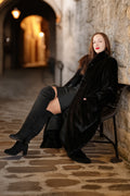 Woman in a black coat and boots sitting against a stone wall in a dimly lit alley.