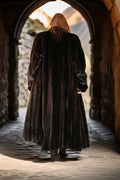 Person in a long dark coat walking through an archway with a stone wall and mountainous background.