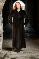 Woman wearing a long dark fur coat standing in a stone archway.