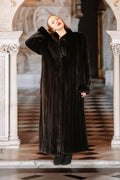 Person wearing a long black coat standing in an elegant interior setting.