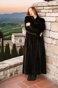 Woman in a long black coat standing against a stone wall with a scenic background