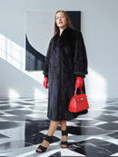 Woman in a black fur coat with red gloves and handbag standing in a modern interior setting.