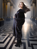 Woman in a dark fur coat standing in an elegant hallway with columns and a bright light source.