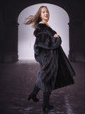 Woman wearing a long black fur coat standing in a dark, archway-like setting.