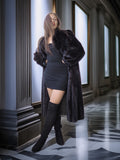 Woman in a long dark fur coat and black knee-high boots standing in an elegant hallway.