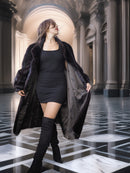 Woman in a long black coat and knee-high boots standing in an elegant interior setting.