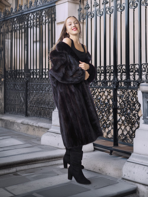 Woman in a long black fur coat standing in front of an ornate black metal gate.