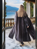 Person wearing a long dark coat standing on a balcony with a scenic view of water and mountains.
