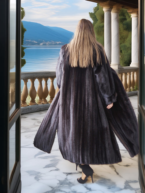 Person wearing a long dark coat standing on a balcony with a scenic view of water and mountains.